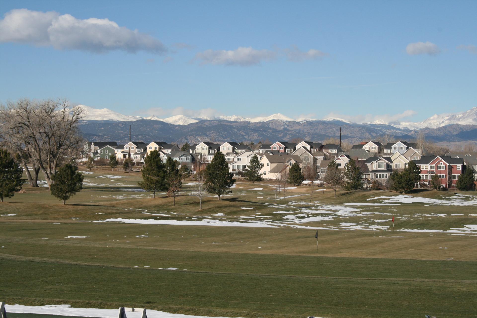 Houses nestled against snowy mountains, with a green field and patches of snow.