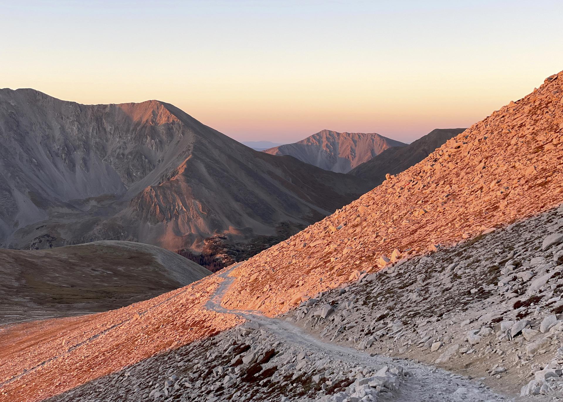 A winding rocky trail ascends barren mountains at golden hour.