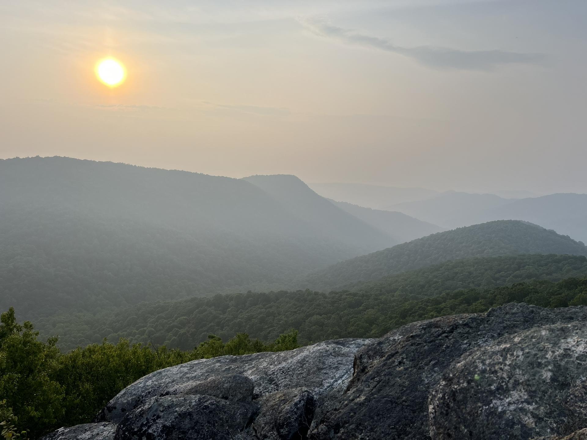 Golden sun over misty forested mountains with prominent rocks in the foreground.