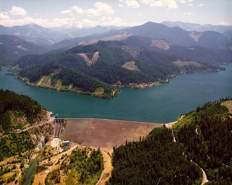 Aerial view of a large dam, reservoir, and forested mountains on a clear day.