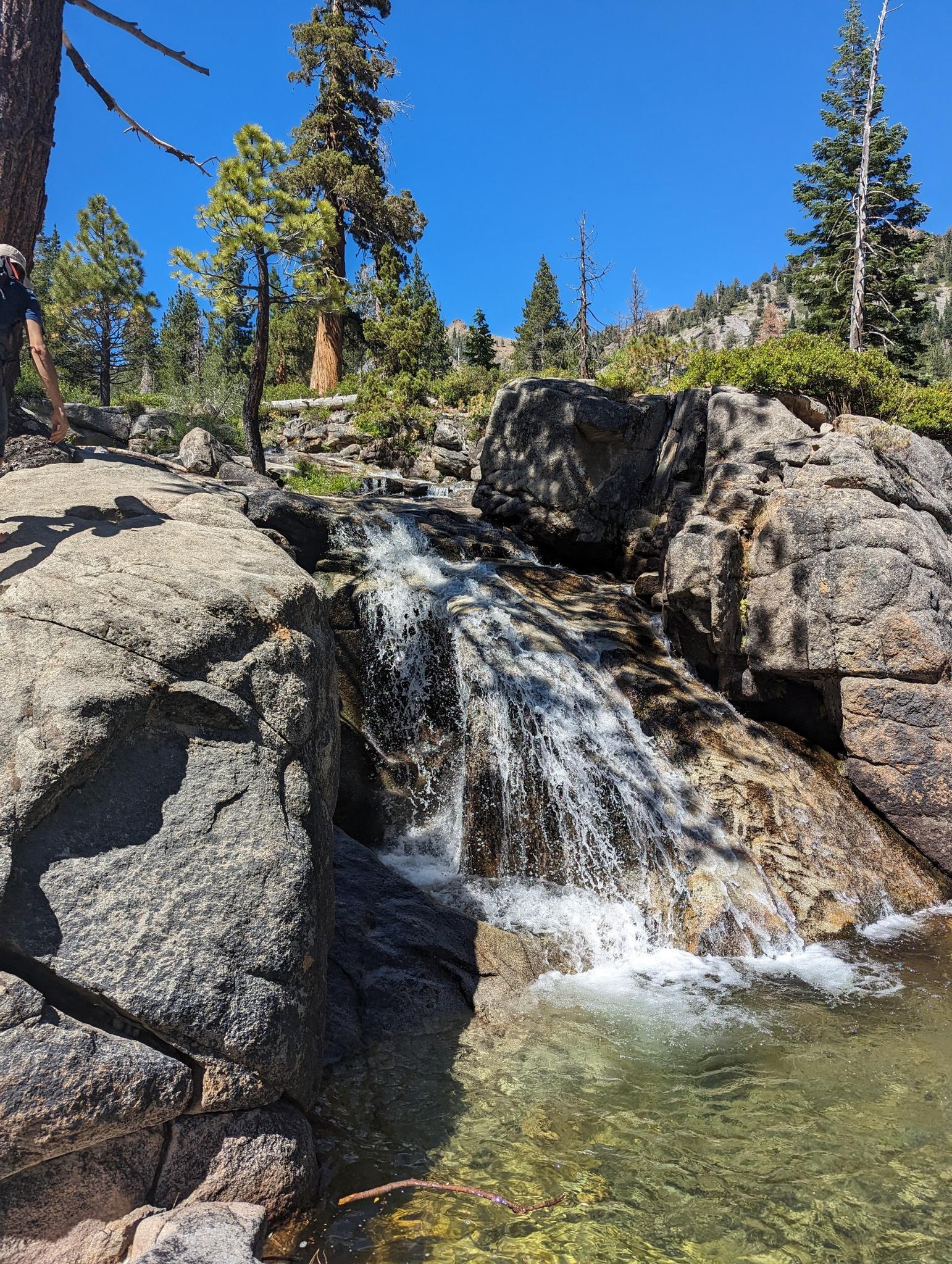 Small waterfall cascading over rocks into a clear pool, bright day.