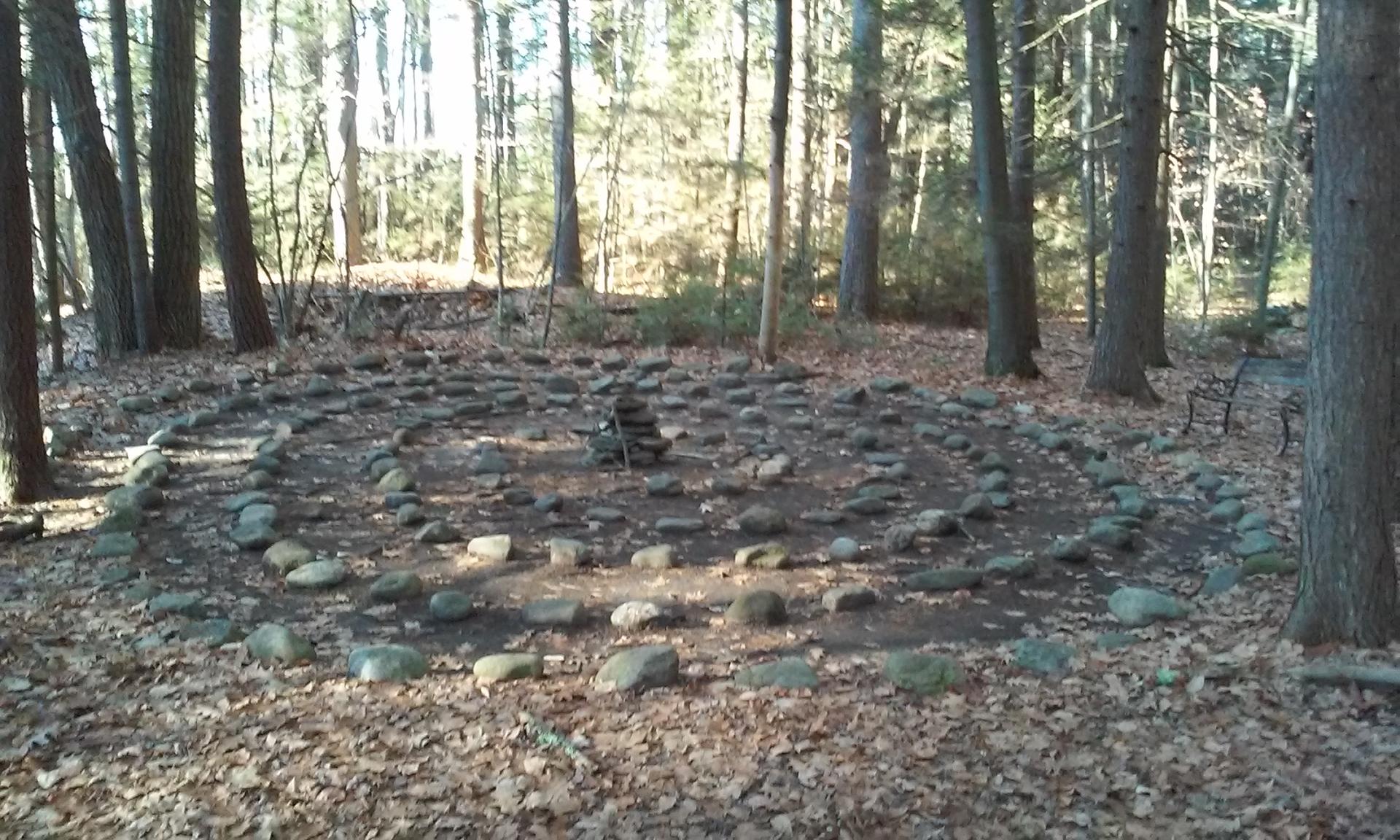 A circular stone labyrinth on a leaf-covered forest floor, surrounded by trees.