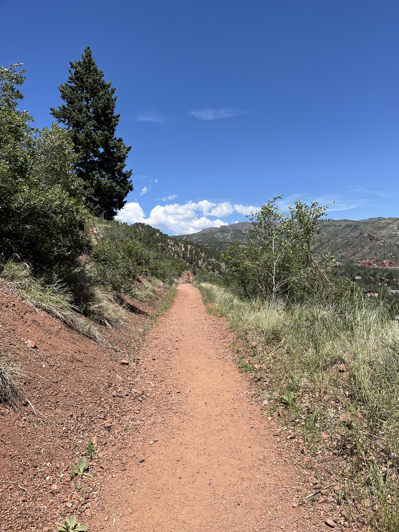 A sunny dirt trail winds through green hills under a bright blue sky.