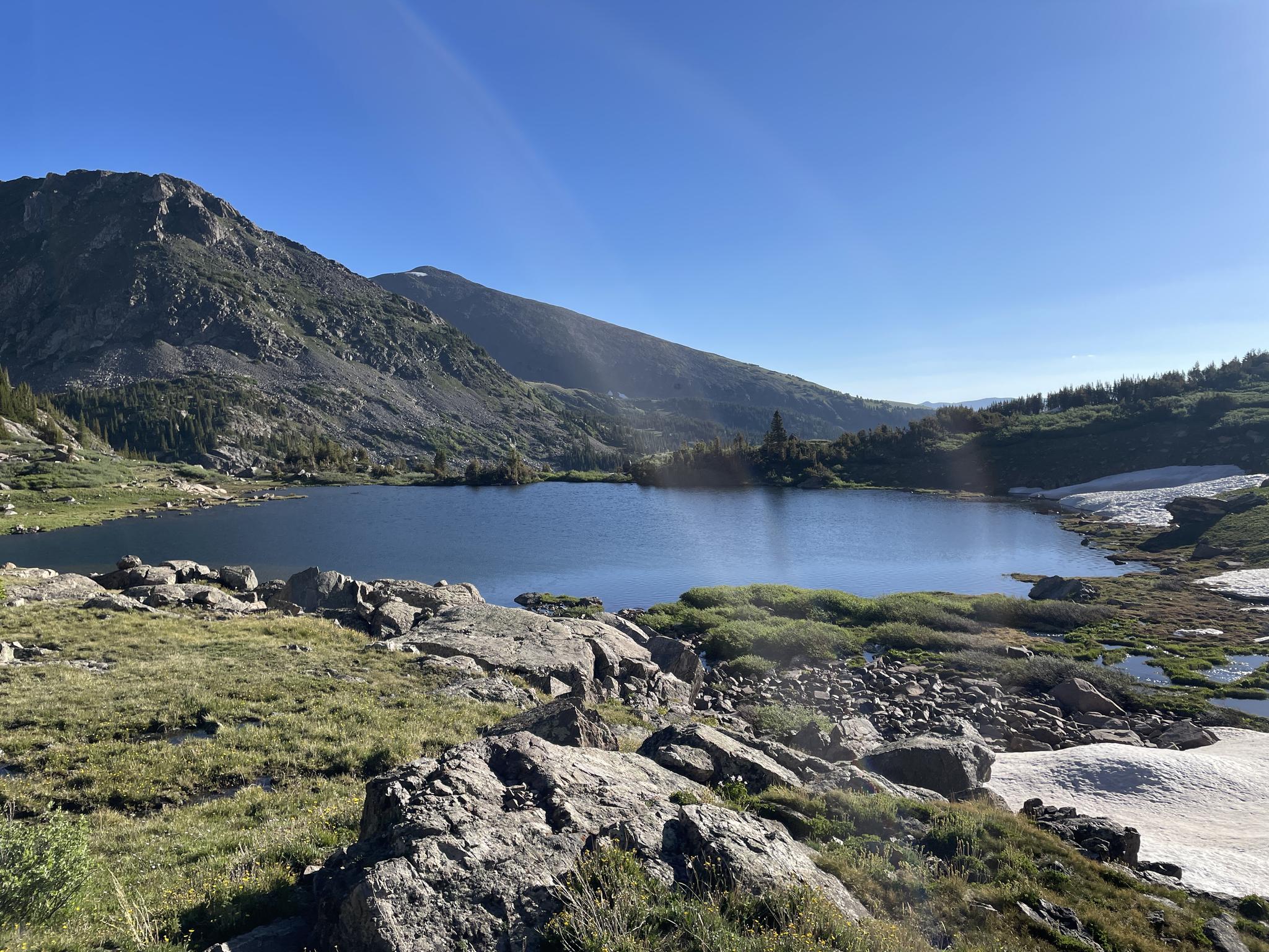 Serene mountain lake with rocky shores, green hills, and snow under blue sky.