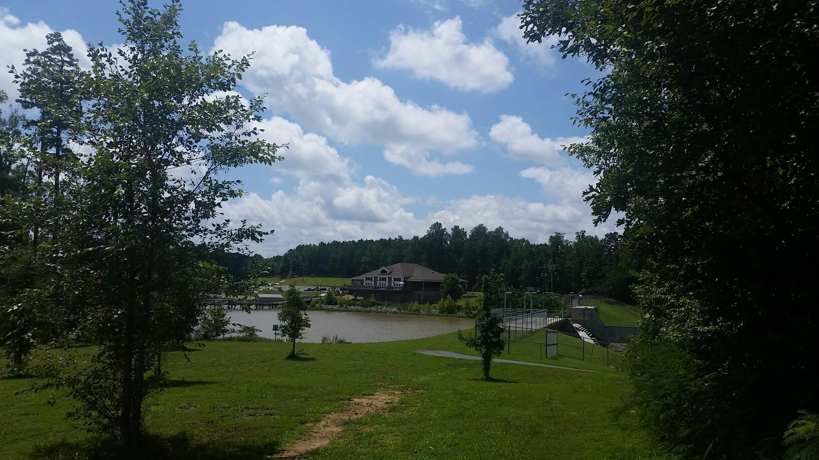 A grassy field overlooks a lake with distant buildings under a cloudy sky.