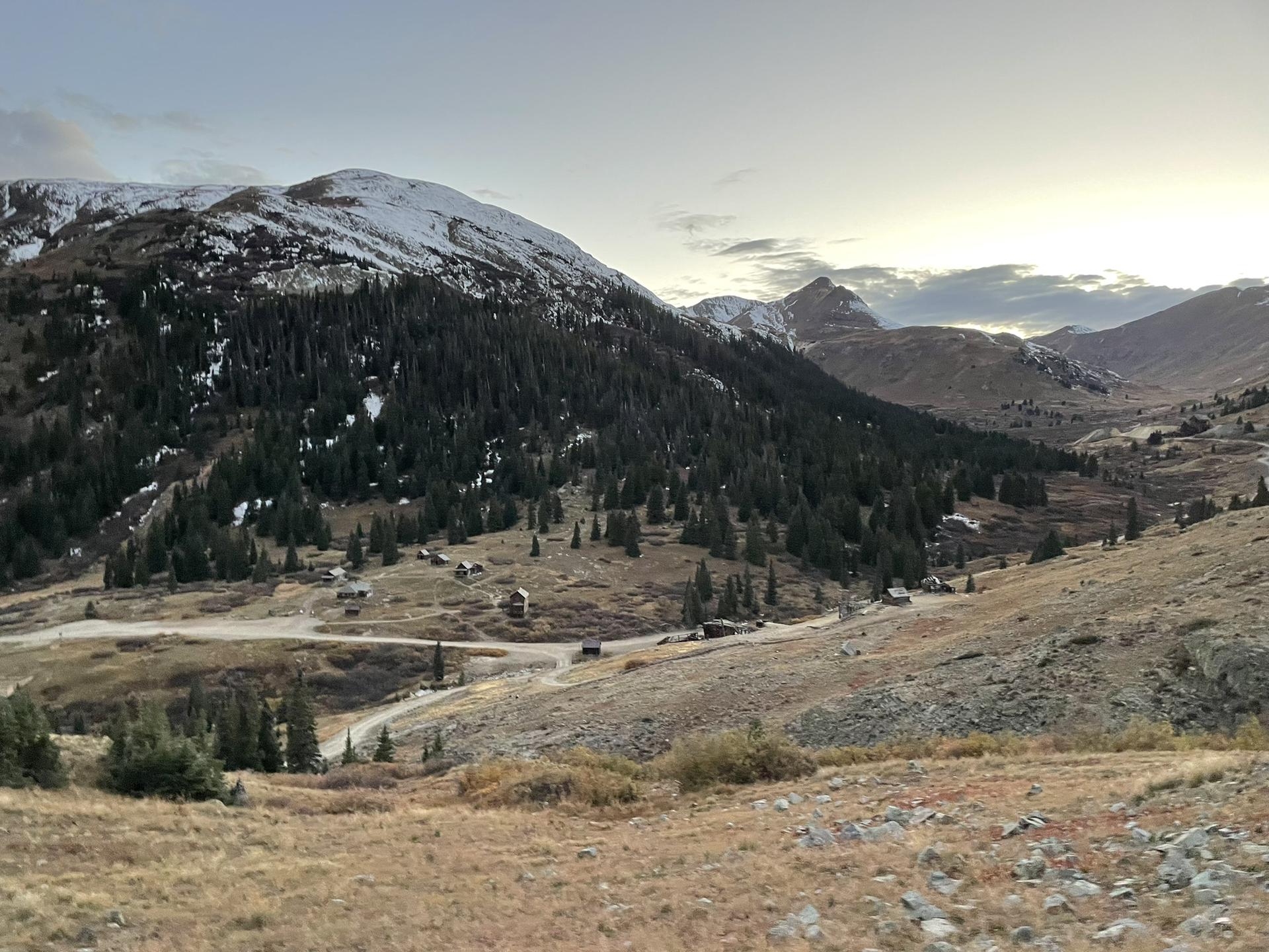 Mountain valley with a winding road, evergreen trees, and snow-capped peaks.