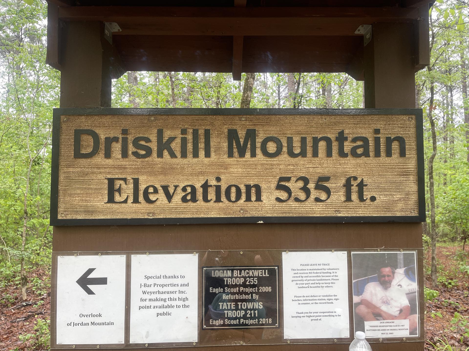 Trailhead sign with elevation information surrounded by green trees under diffused light.