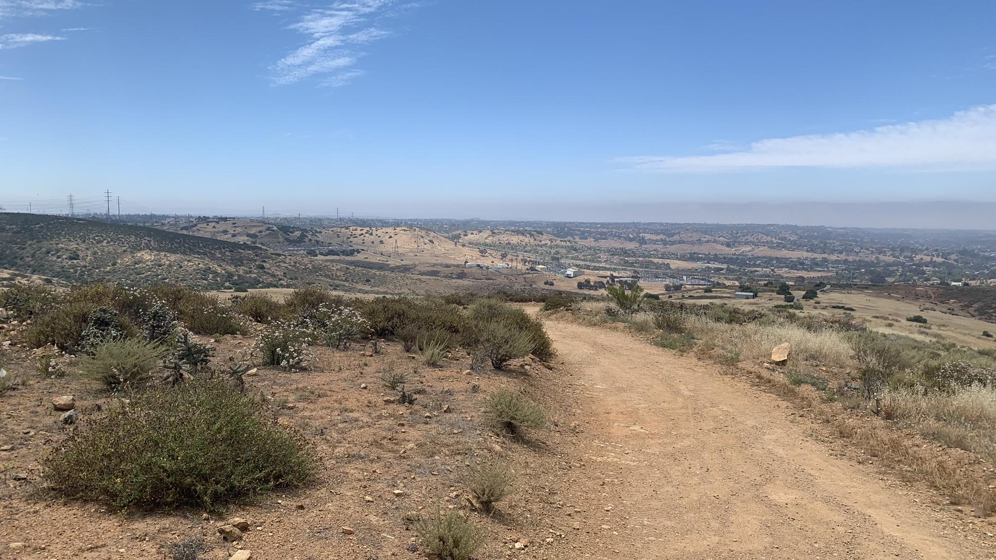 A winding dirt trail through dry, brushy hills under a clear blue sky.