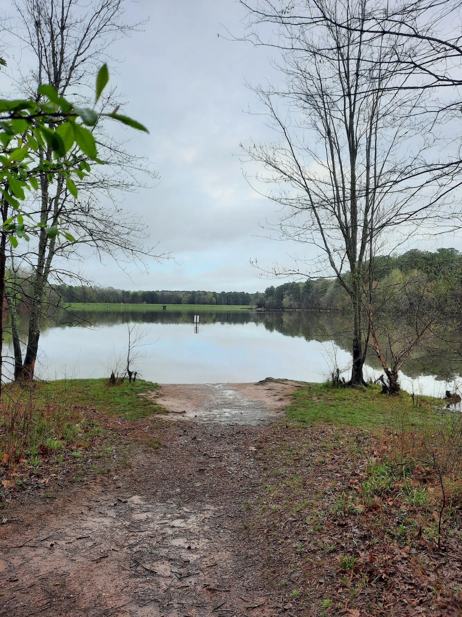 A dirt path leads to a calm lake surrounded by bare trees under a cloudy sky.