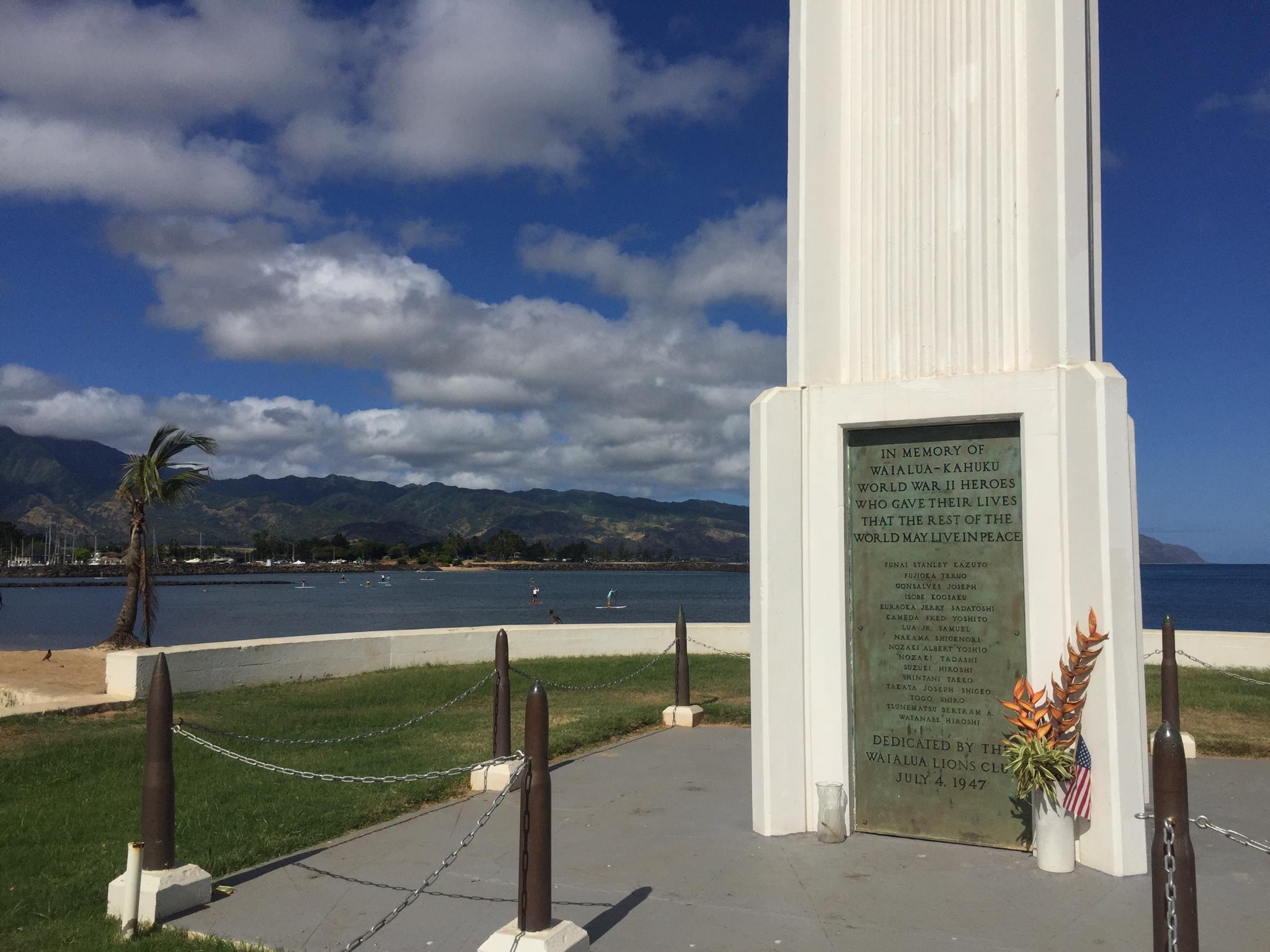 White monument with plaque by the coast, mountains in background, sunny day.