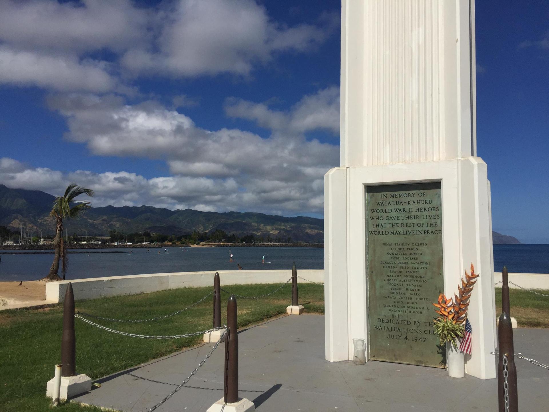 White monument with plaque by the coast, mountains in background, sunny day.
