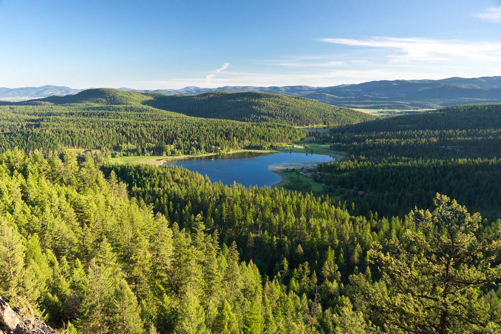 A serene blue lake nestled within a vast, sunlit green forest and hills.