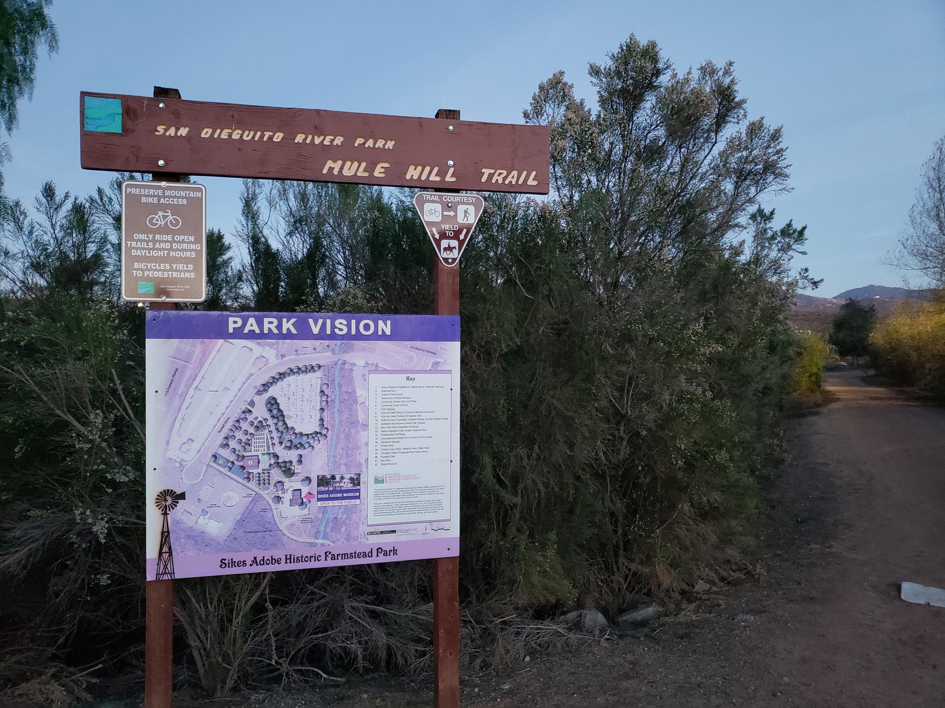 A wooden trail sign and park map stand beside a dirt path.