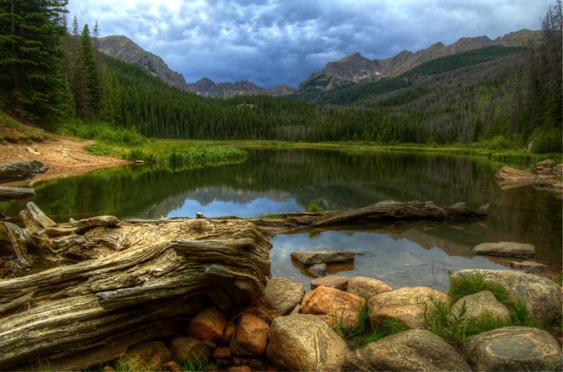 Overcast sky above a serene mountain lake, surrounded by forests and peaks.