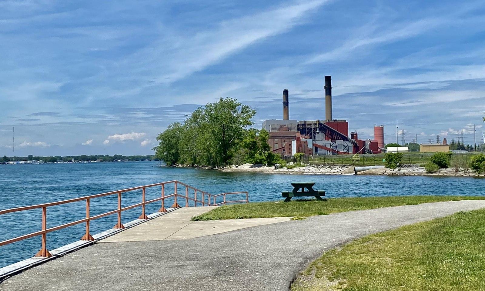 Industrial building with smokestacks across water, with a park path and picnic table.
