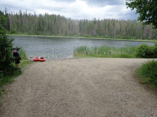 Person by a lake with two kayaks, dense forest under cloudy sky.