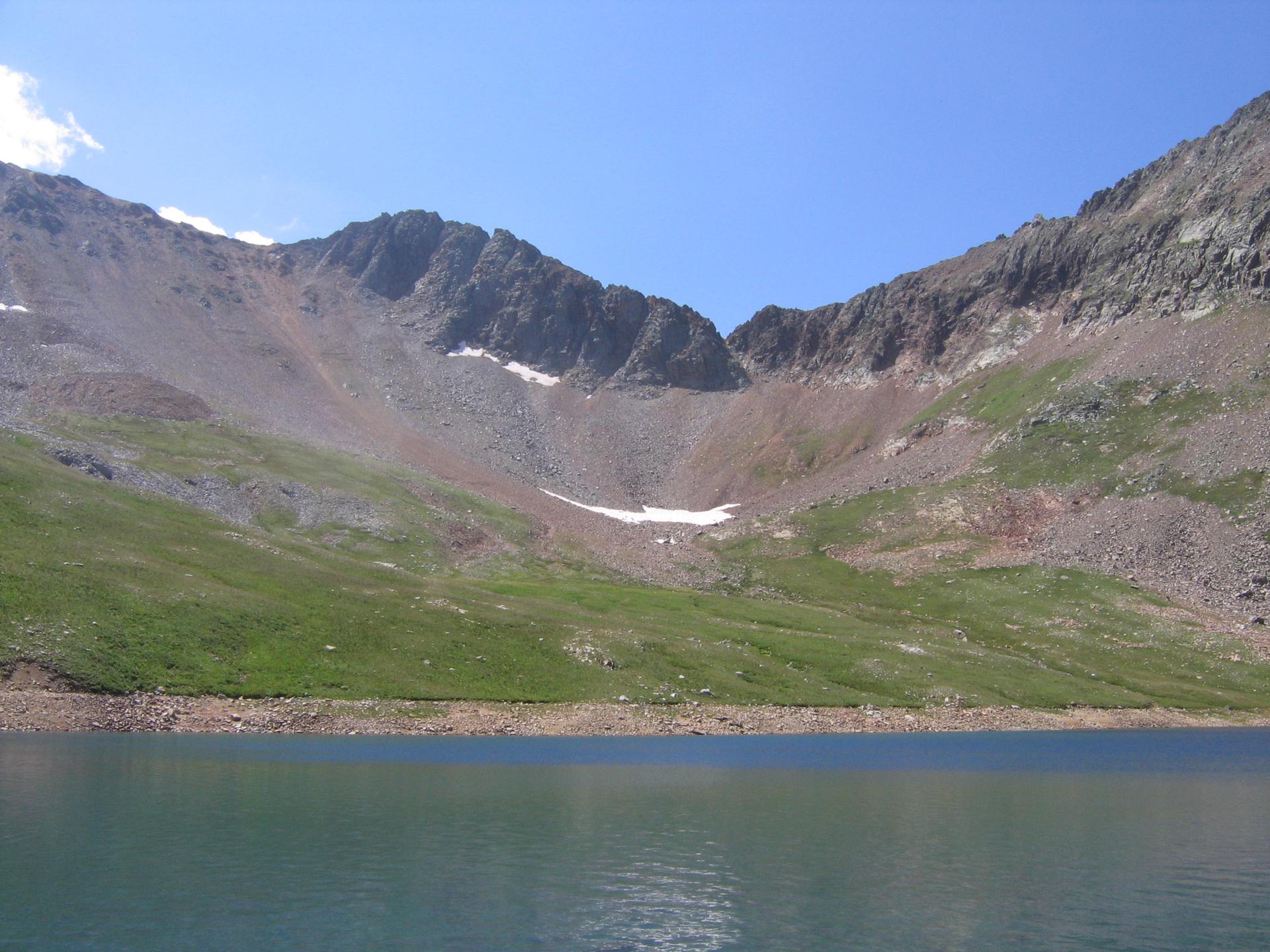 Brightly lit mountain lake with rocky, green slopes and patches of snow.
