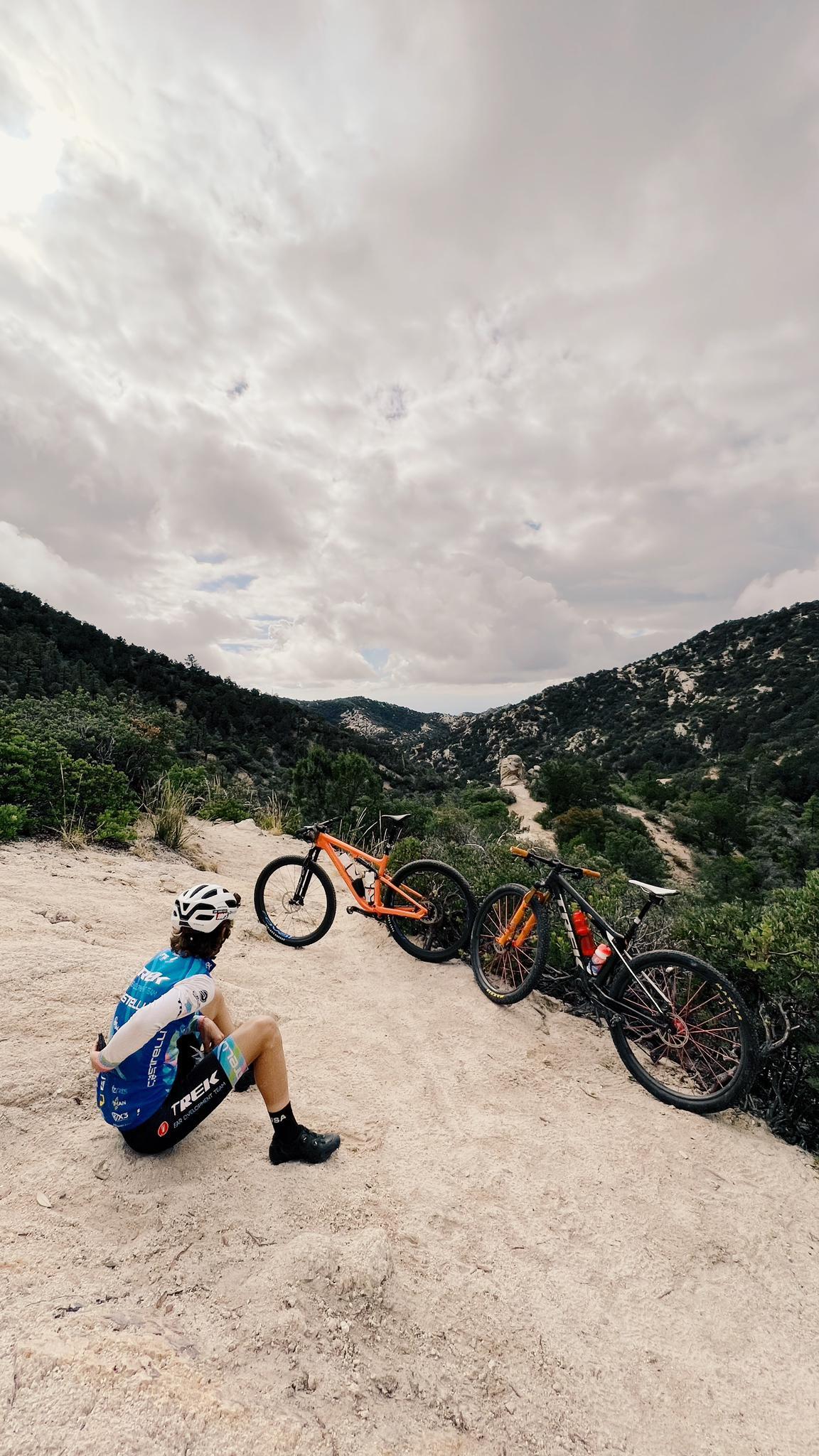 Biker rests on a dirt trail next to two mountain bikes under cloudy skies.