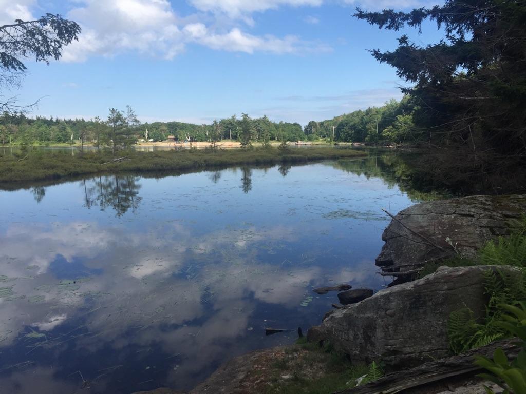 Calm water reflects a blue sky with clouds, surrounded by trees and rocks.