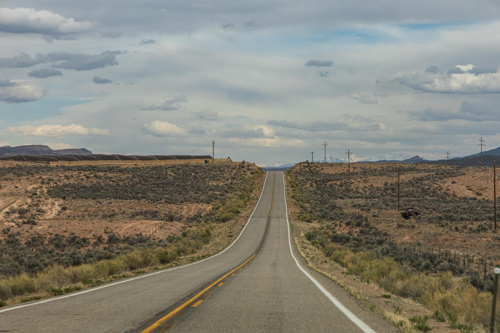 A long, straight road ascends through a dry, hilly landscape under a cloudy sky.