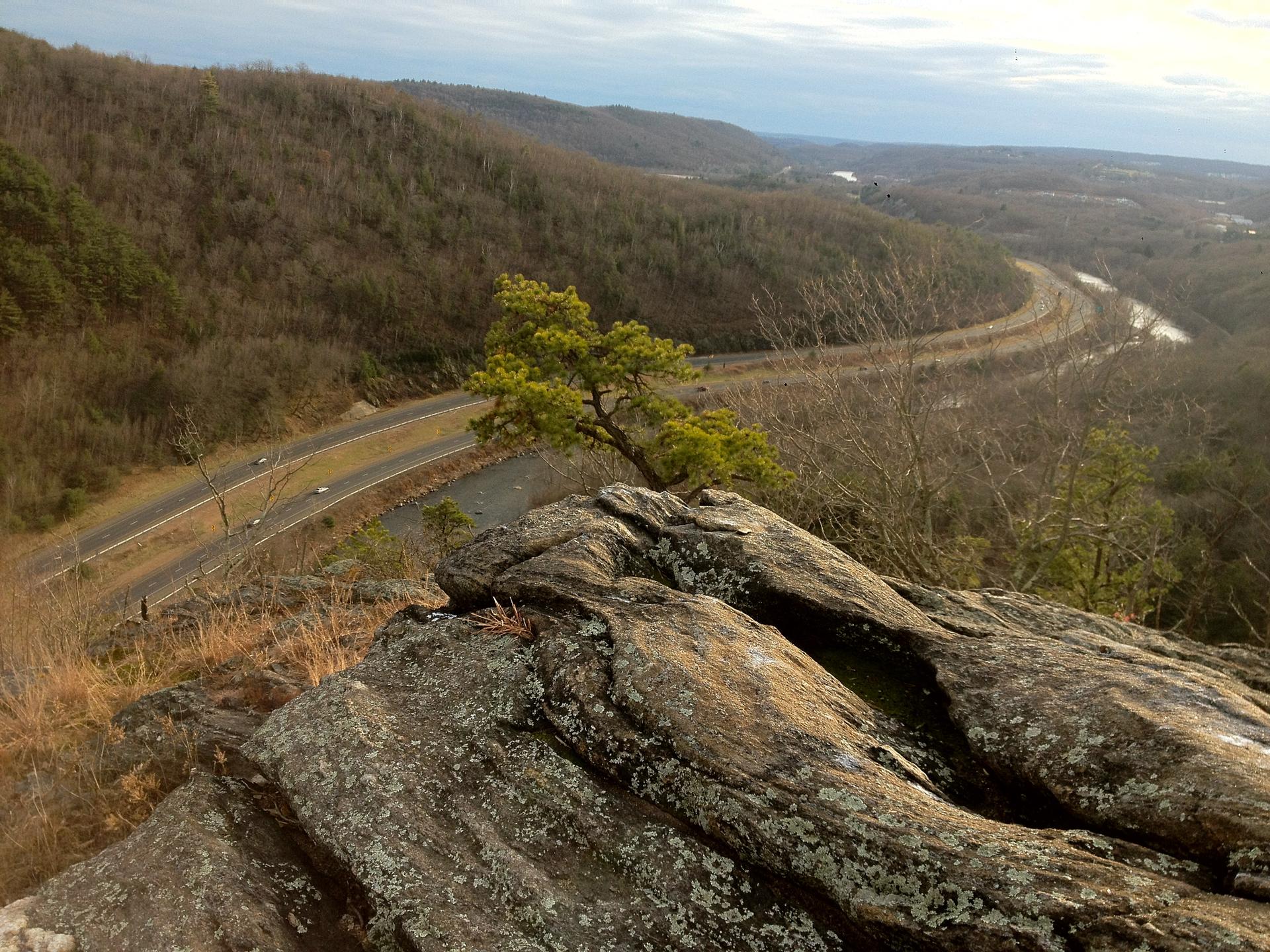 Rocky outcrop overlooks a winding road, river, and forested hills under diffused light.