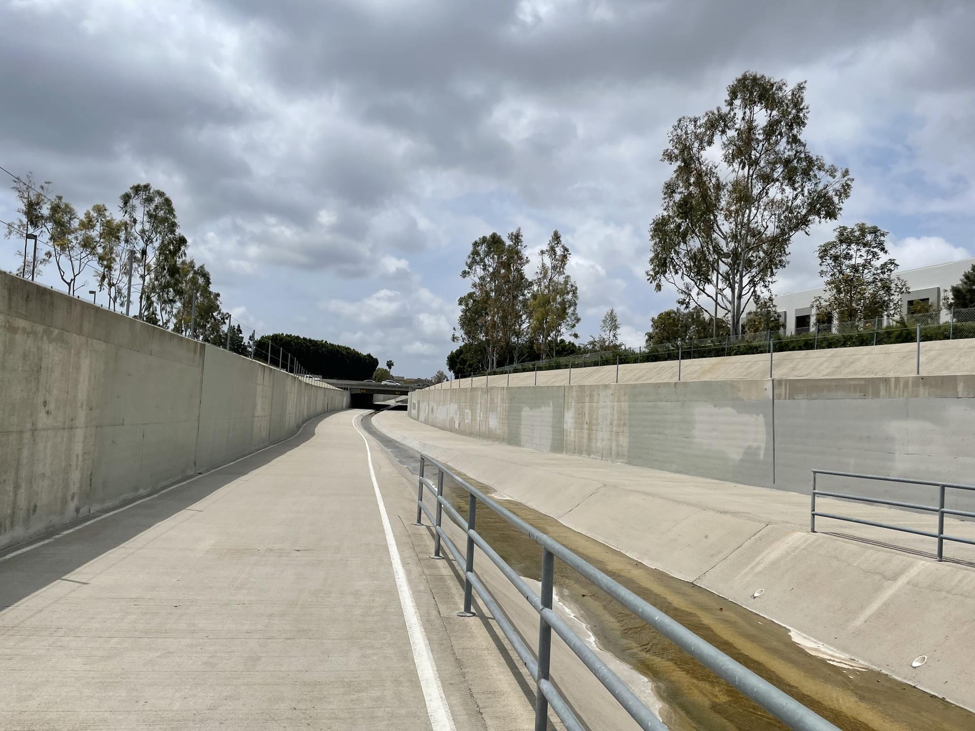 Concrete channel with a paved path, metal railing, and trees under cloudy sky.