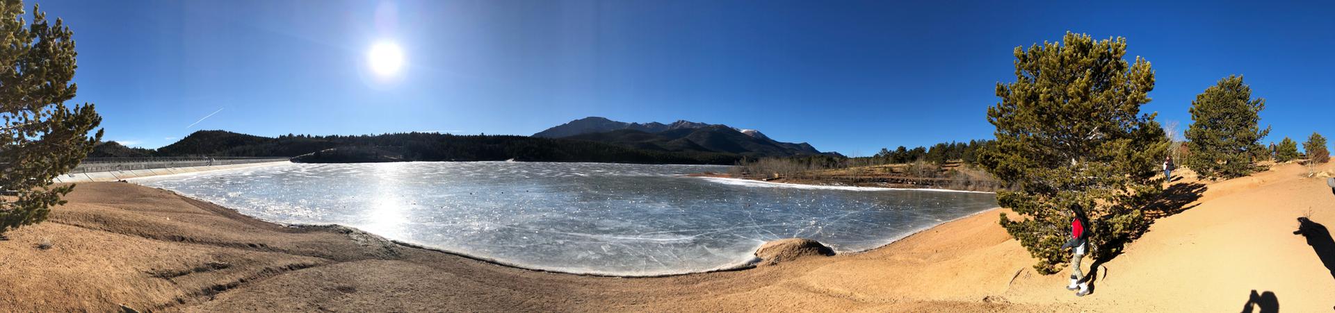 Bright panoramic view of a partially frozen lake, mountains, and pine trees. A person rests.