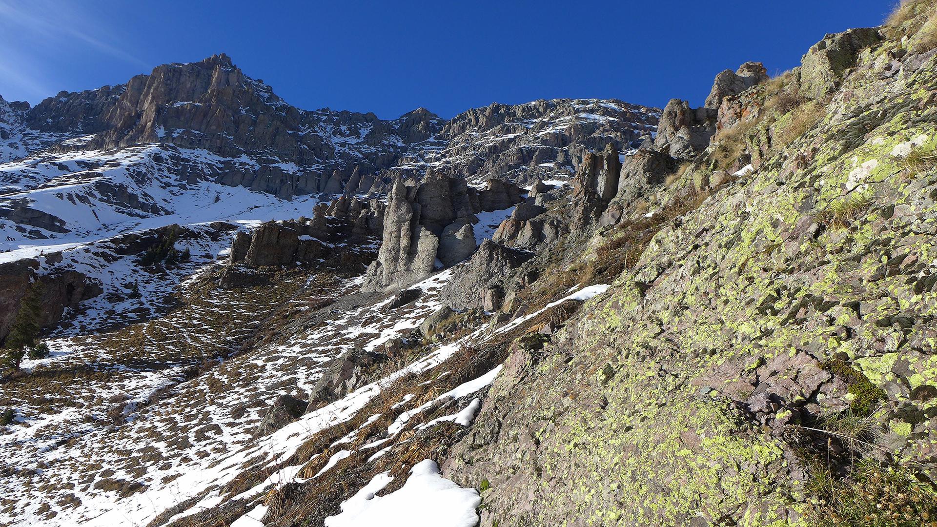 A steep, rocky mountain slope with patches of snow under a clear blue sky.