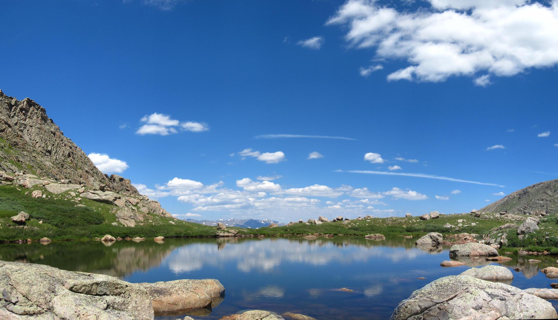 Calm mountain lake reflecting blue sky, surrounded by rocks and green hills.