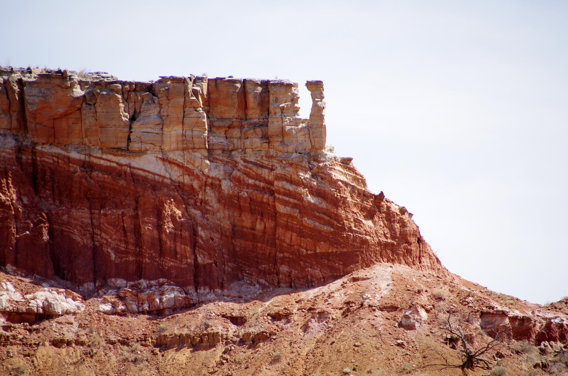 Striated red and white rock cliff face with a unique pinnacle against a bright sky.