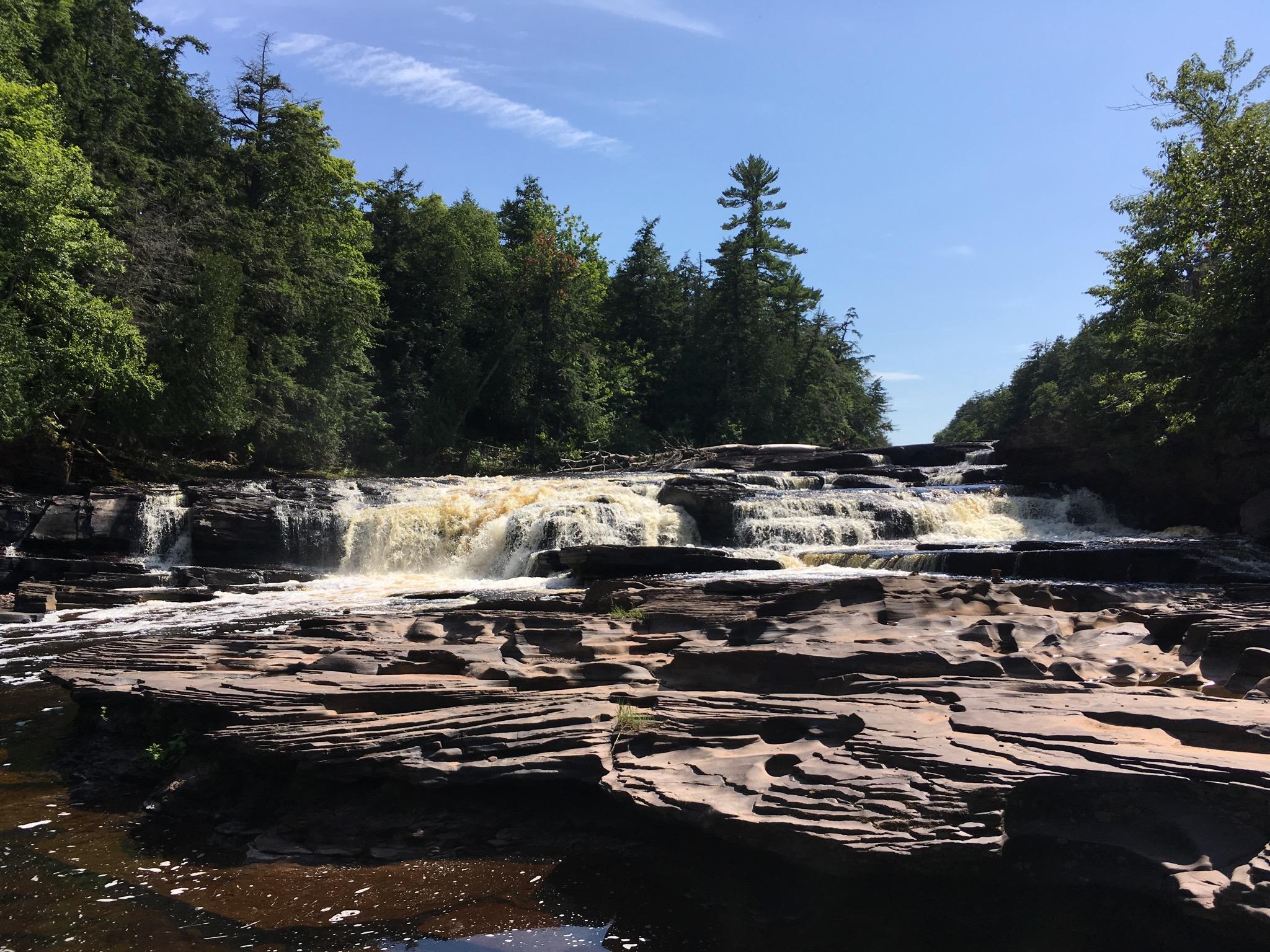 Wide, tiered waterfall cascades over dark rocks under a bright blue sky.