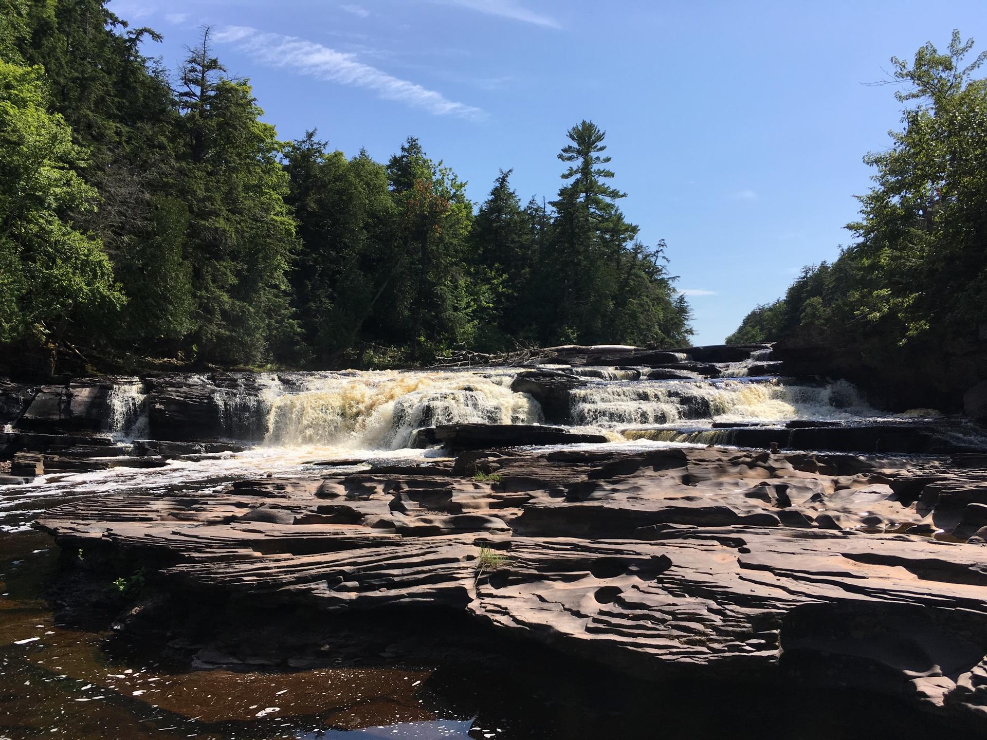 Wide, tiered waterfall cascades over dark rocks under a bright blue sky.