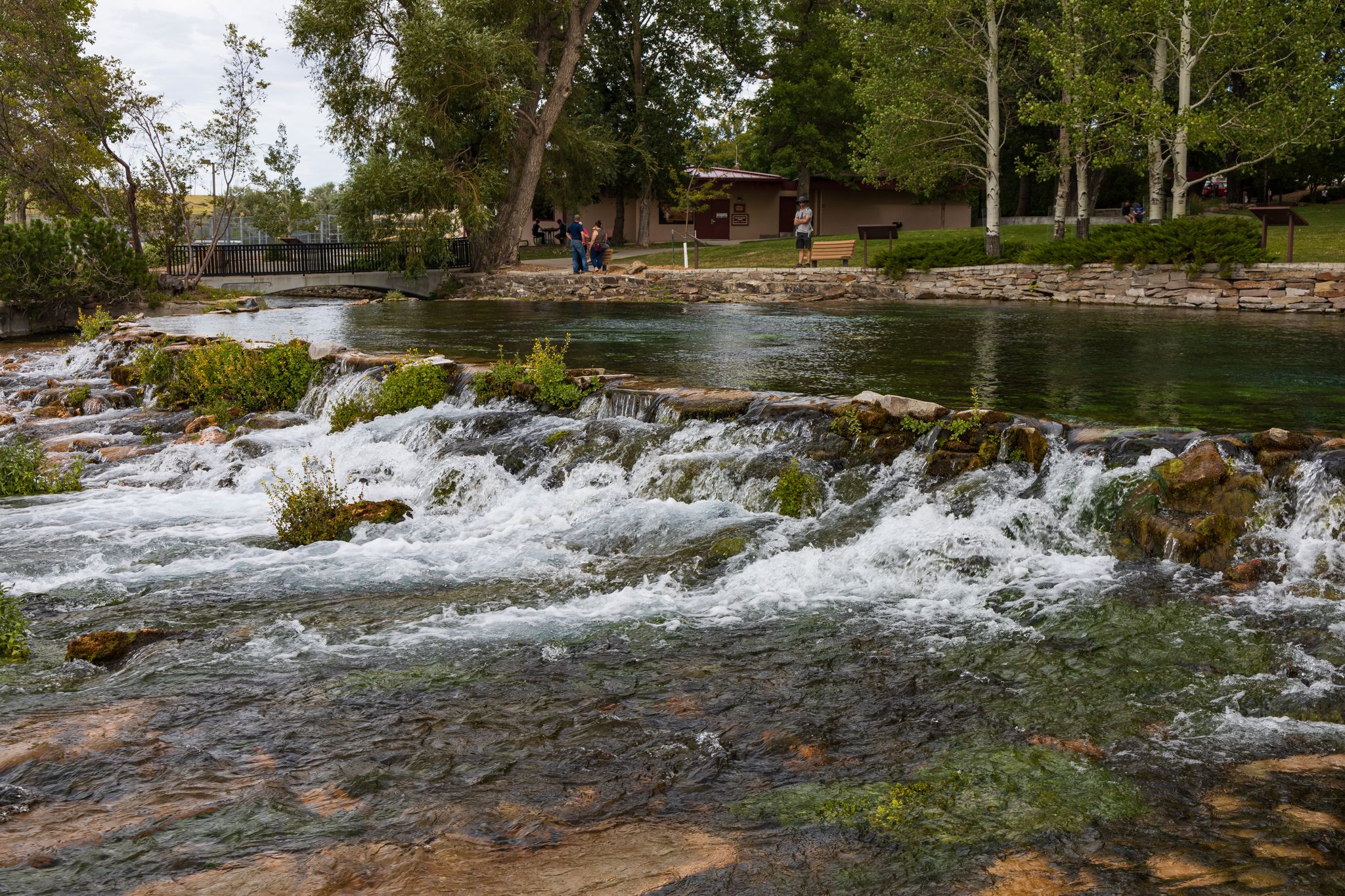 Rushing river with small cascades, green trees, and people on the far bank. - River's Edge Trail