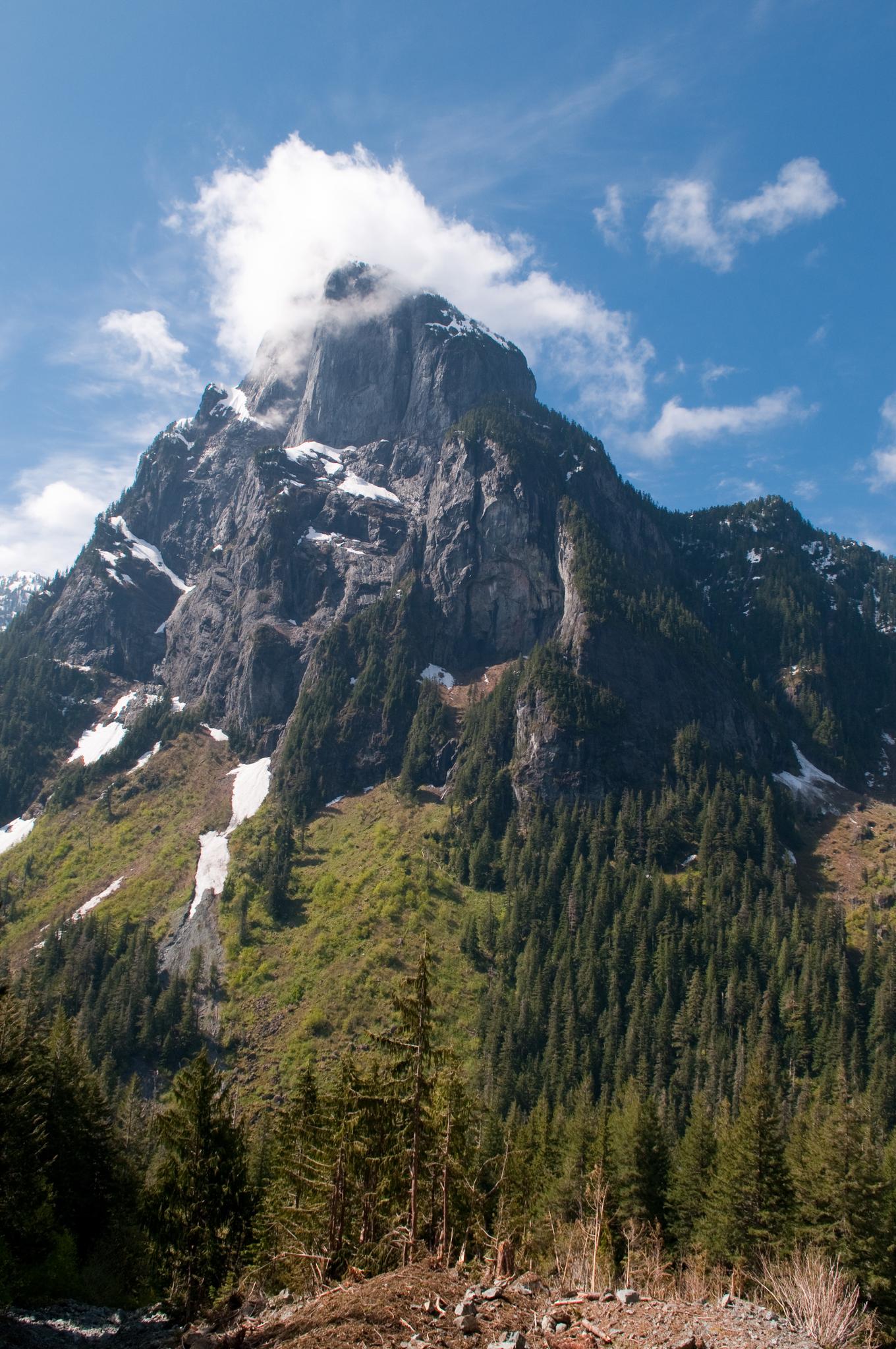 Rugged mountain peak with snow patches, green trees, and a cloud-capped summit.