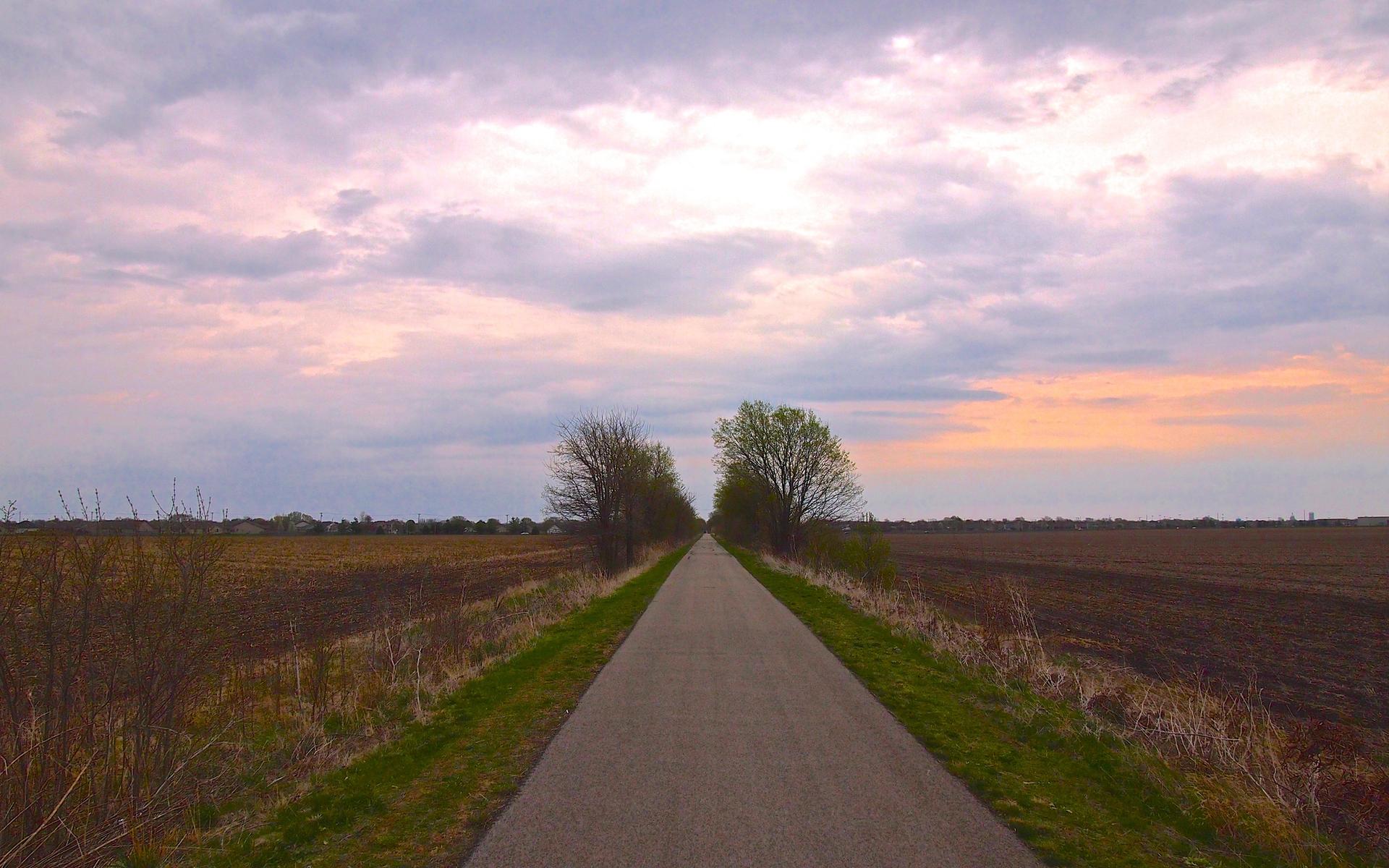 A straight paved path extends through fields under a dramatic, colorful sky.