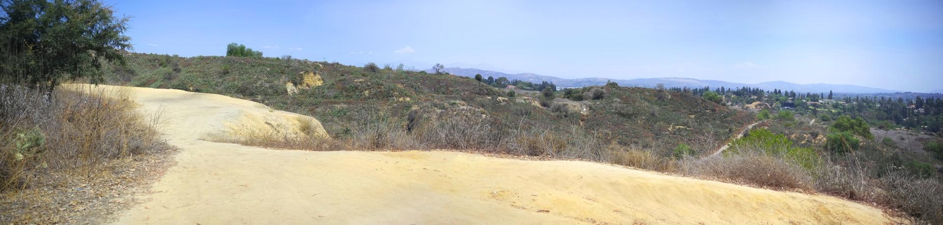 A wide dirt path winds through dry, hilly scrubland under a bright blue sky.