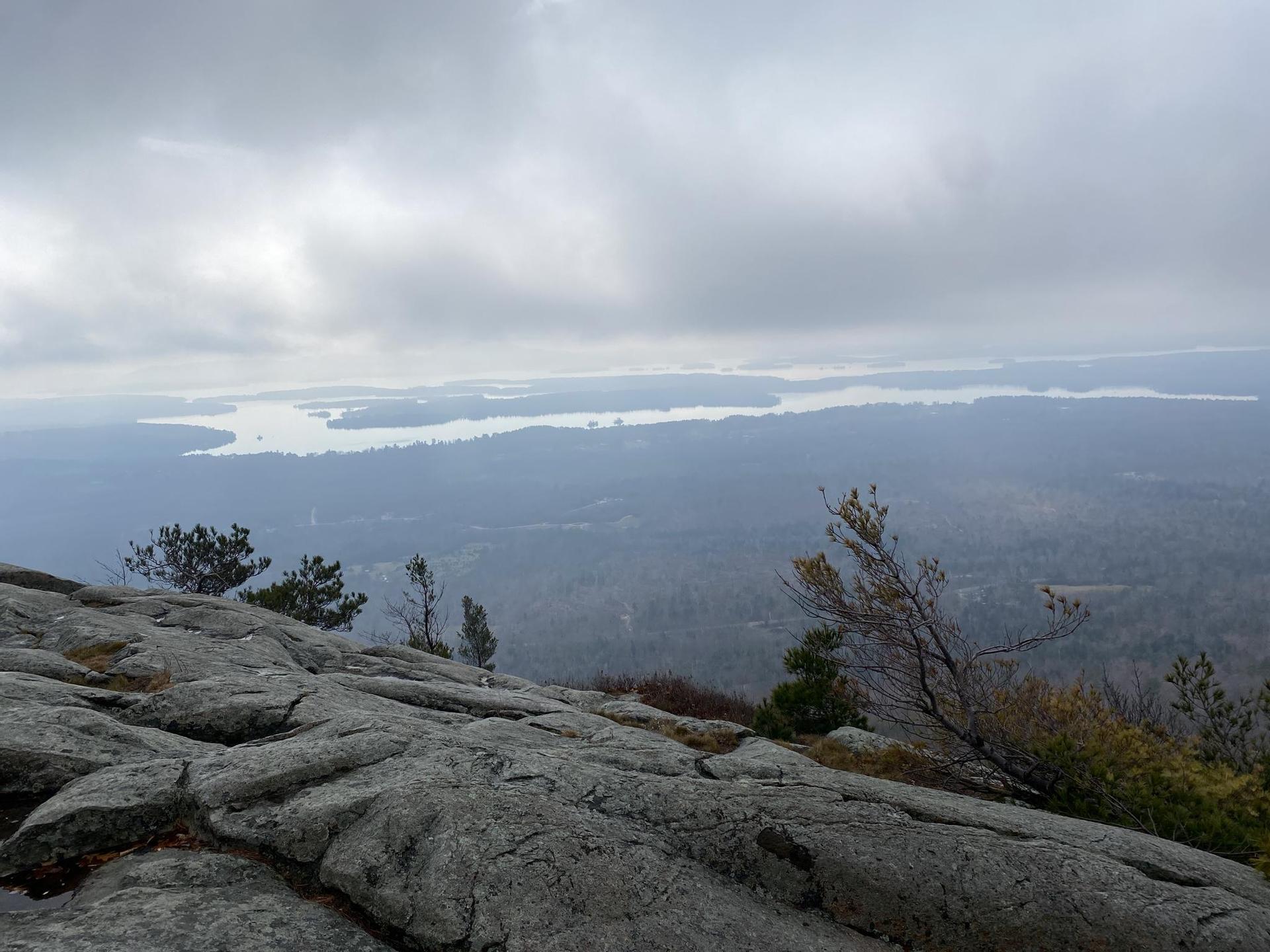 Rocky outcrop overlooking a vast forested landscape and distant water under overcast skies.
