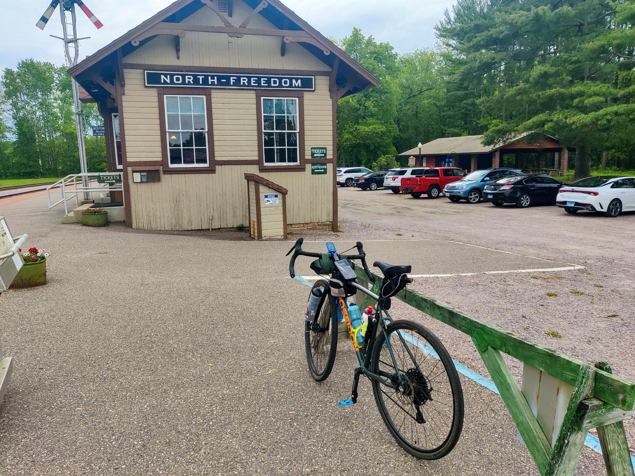 Bicycle parked on gravel near a small building with cars in background.