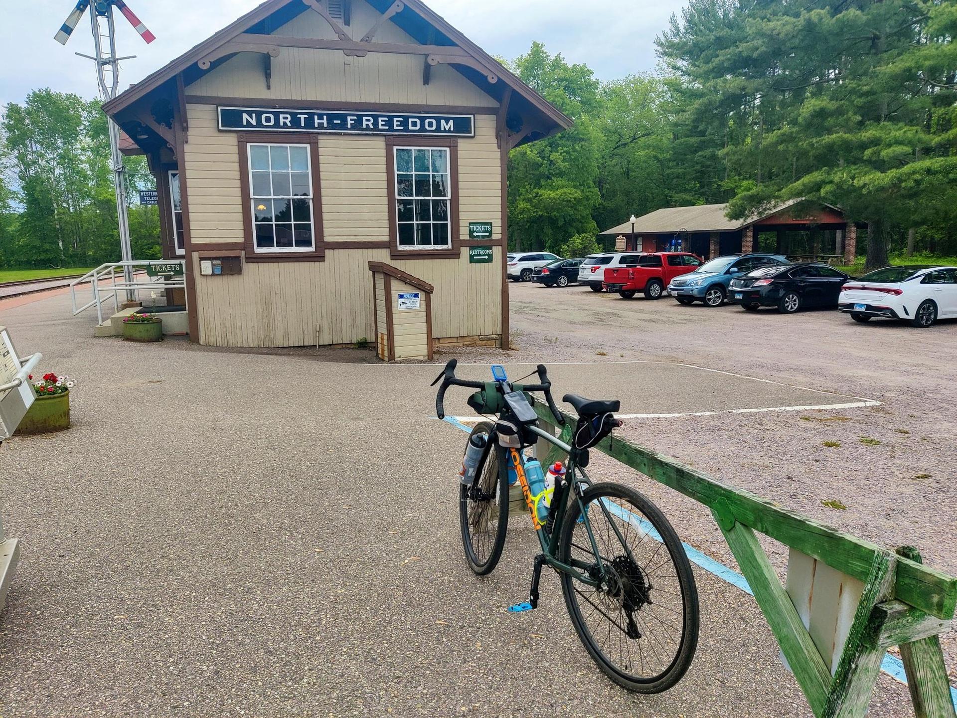 Bicycle parked on gravel near a small building with cars in background.