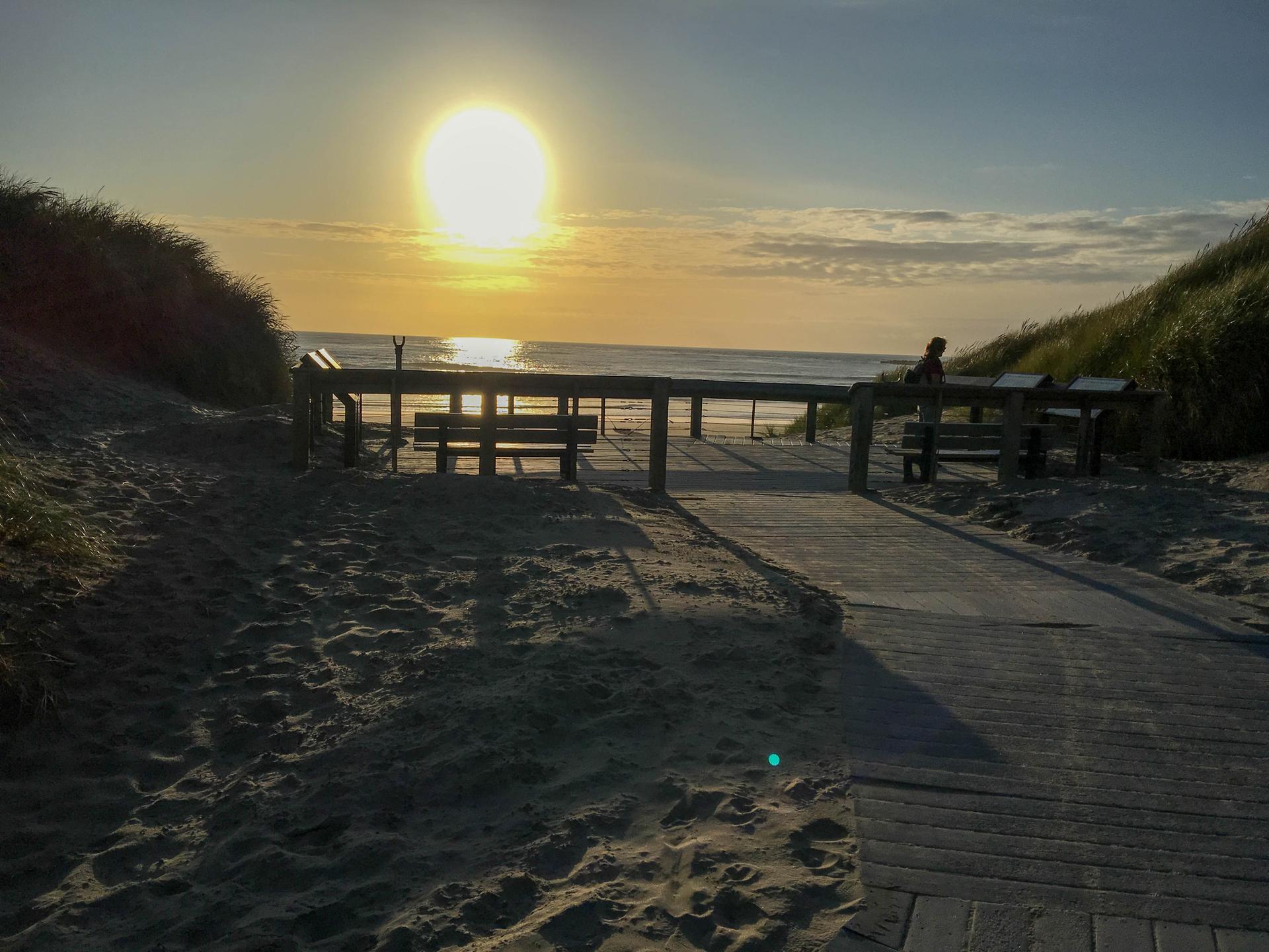 Bright sunset illuminates a beach boardwalk, dunes, and a person viewing the ocean.