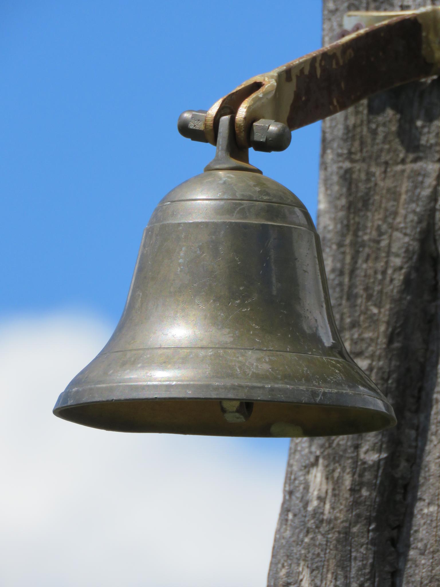 An old brass bell hangs from a wooden post against a bright blue sky.