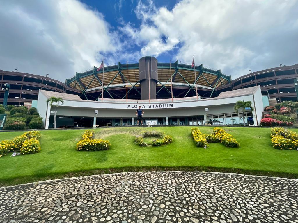 Large stadium building under a partly cloudy sky, with green lawn and flower bushes.