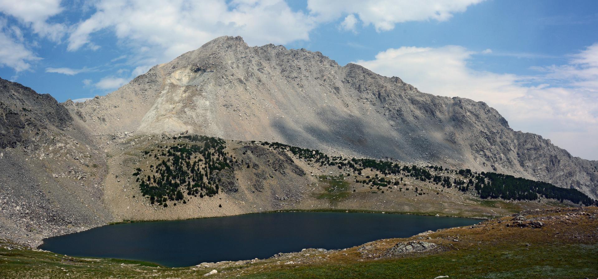 Dark alpine lake nestled below a large rocky mountain under a cloudy sky.