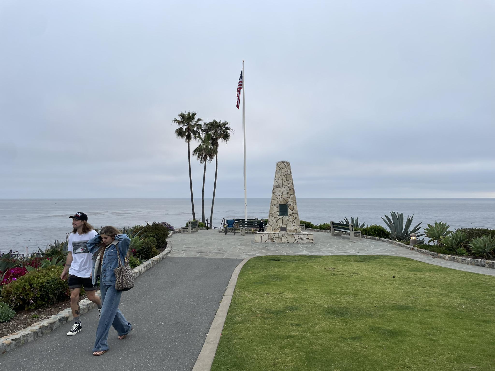 Two people walk past a monument and flag overlooking a cloudy ocean.