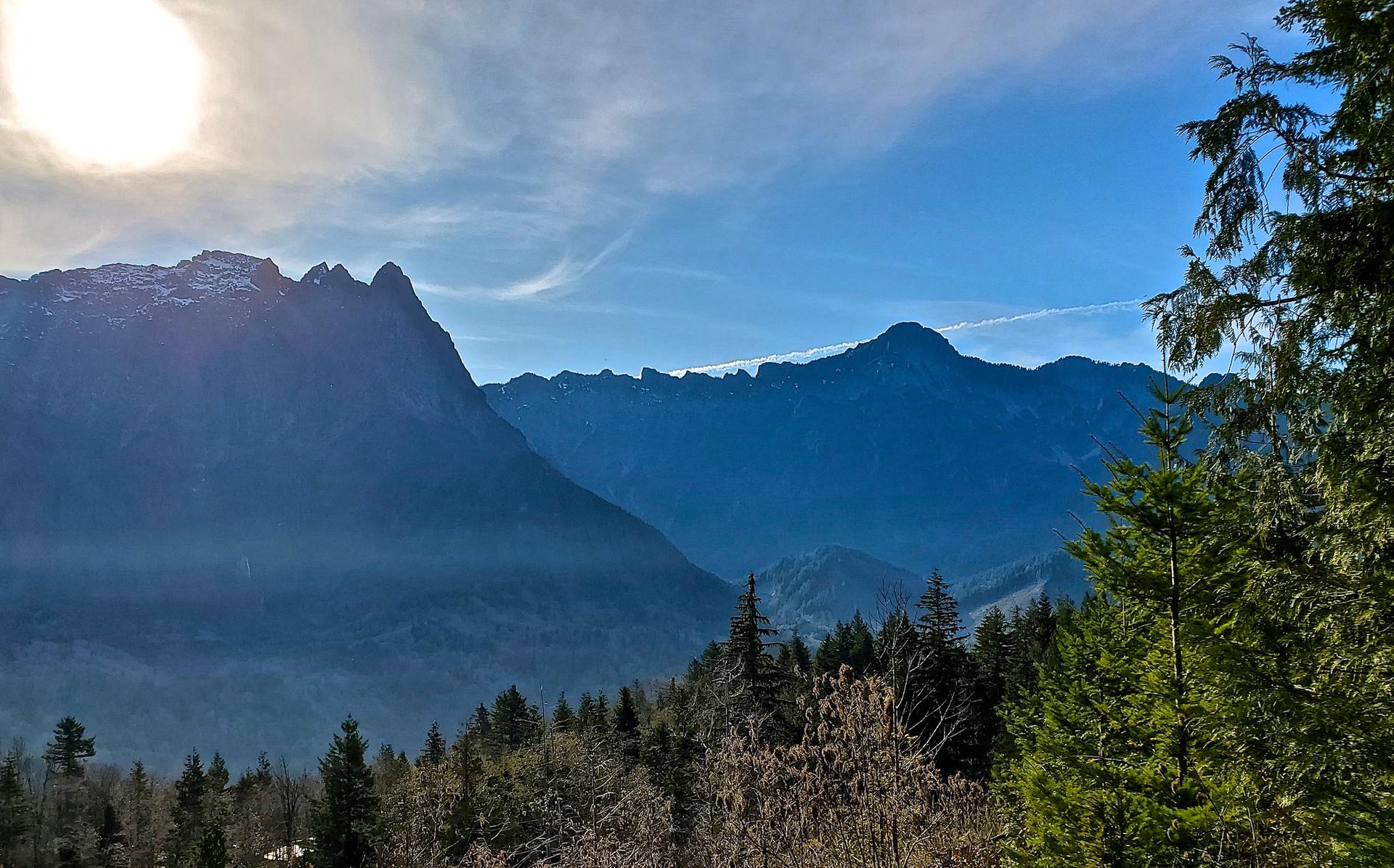 Bright sun over majestic mountains and a forested valley under a blue sky.