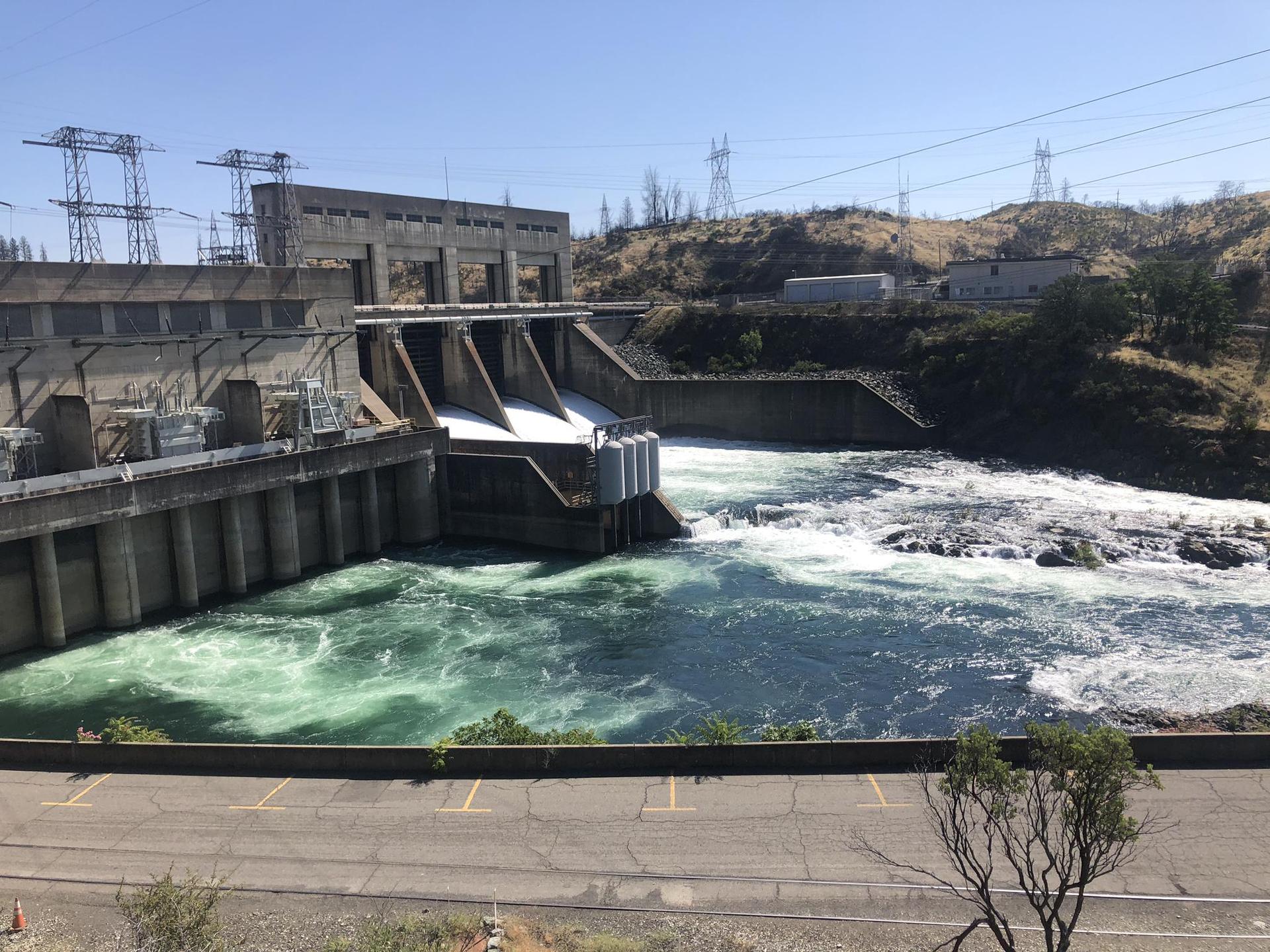 Concrete dam releasing water into a turbulent river on a sunny day.