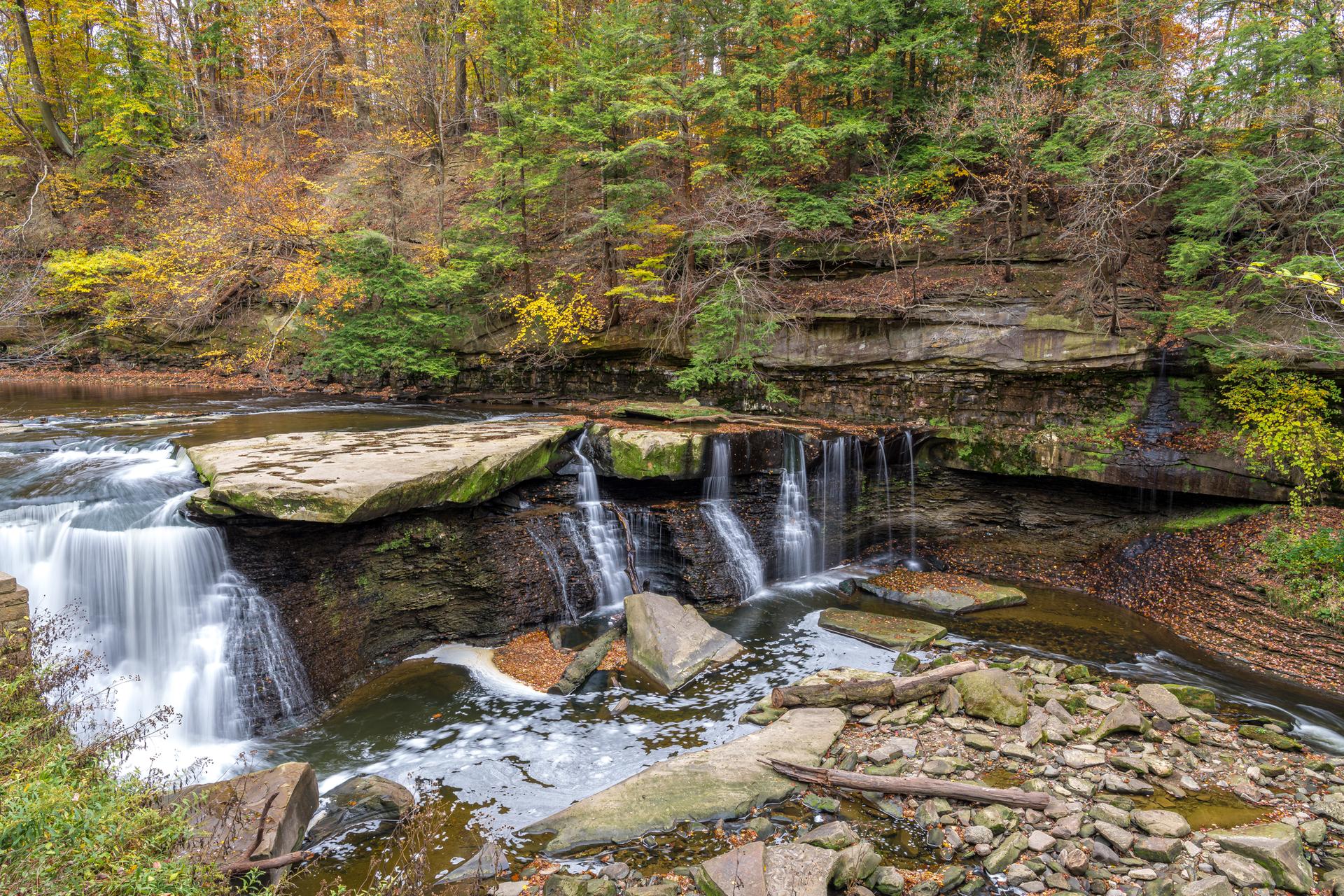 A wide waterfall cascades over rocky ledges in an autumn forest.