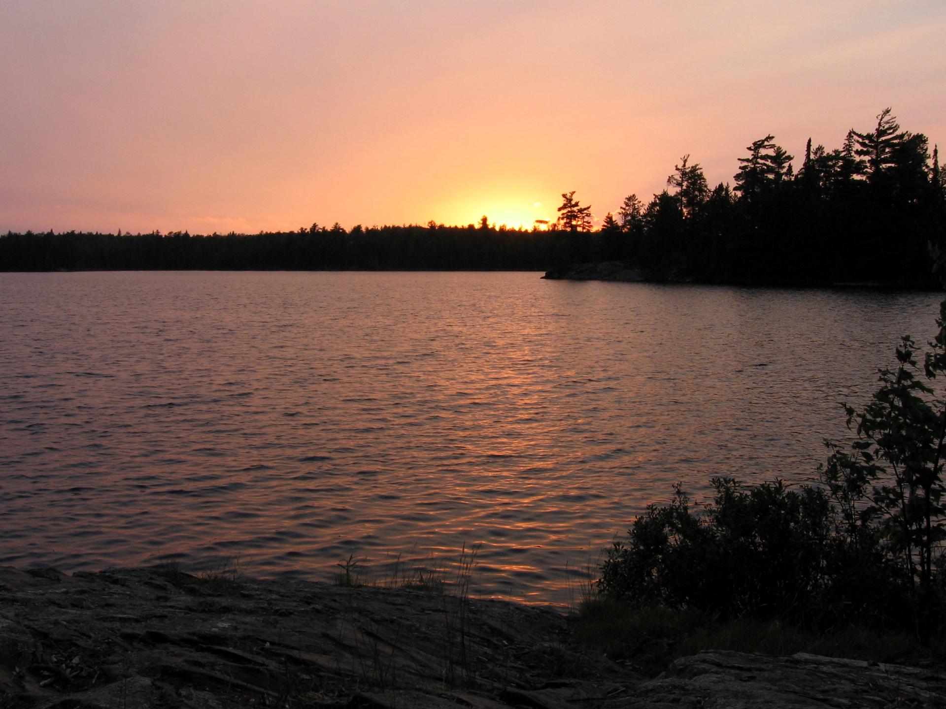Serene lake at sunset with orange sky and dark forested shoreline.