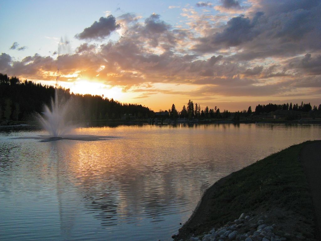Sunset over a lake with a fountain, reflecting golden light on the water. - Riverstone Park Loop