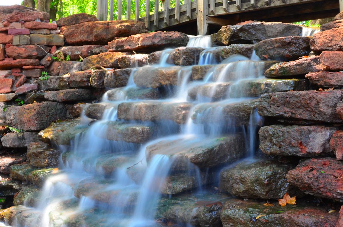 Water cascades over reddish-brown rocks under a wooden bridge in bright light.